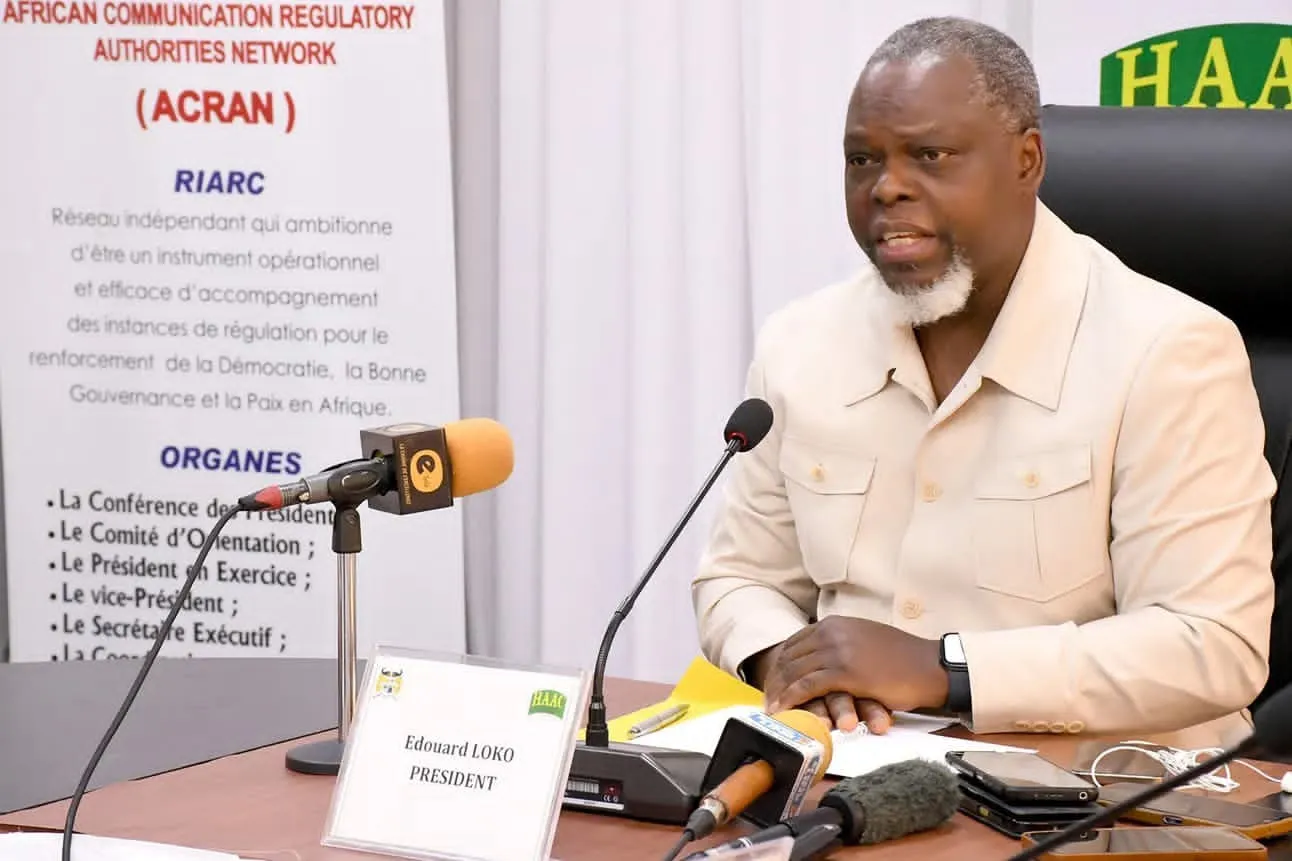 Un homme en veste beige prend la parole devant des micros lors d une réunion officielle