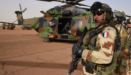 Militaire en tenue de camouflage tenant une arme devant un hélicoptère de transport militaire au sol