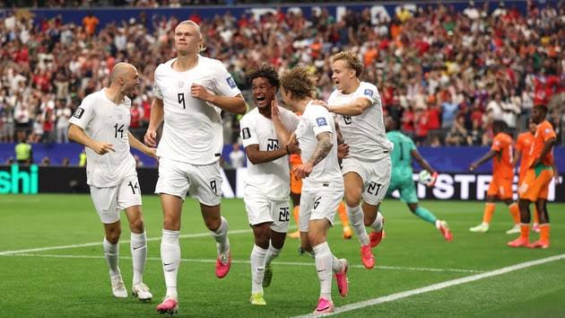 Joueurs en blanc célébrant un but lors d’un match de football devant des supporters en tribunes et adversaires en orange.