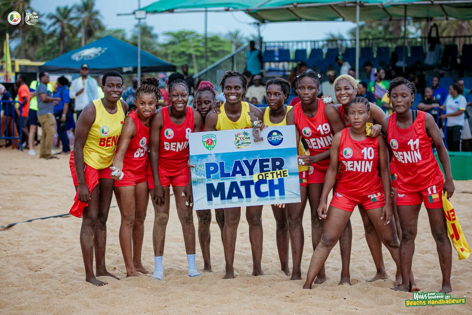 Dix joueuses de beach handball posent ensemble sur le sable avec un panneau « Player of the Match » devant des spectateurs assis et debout