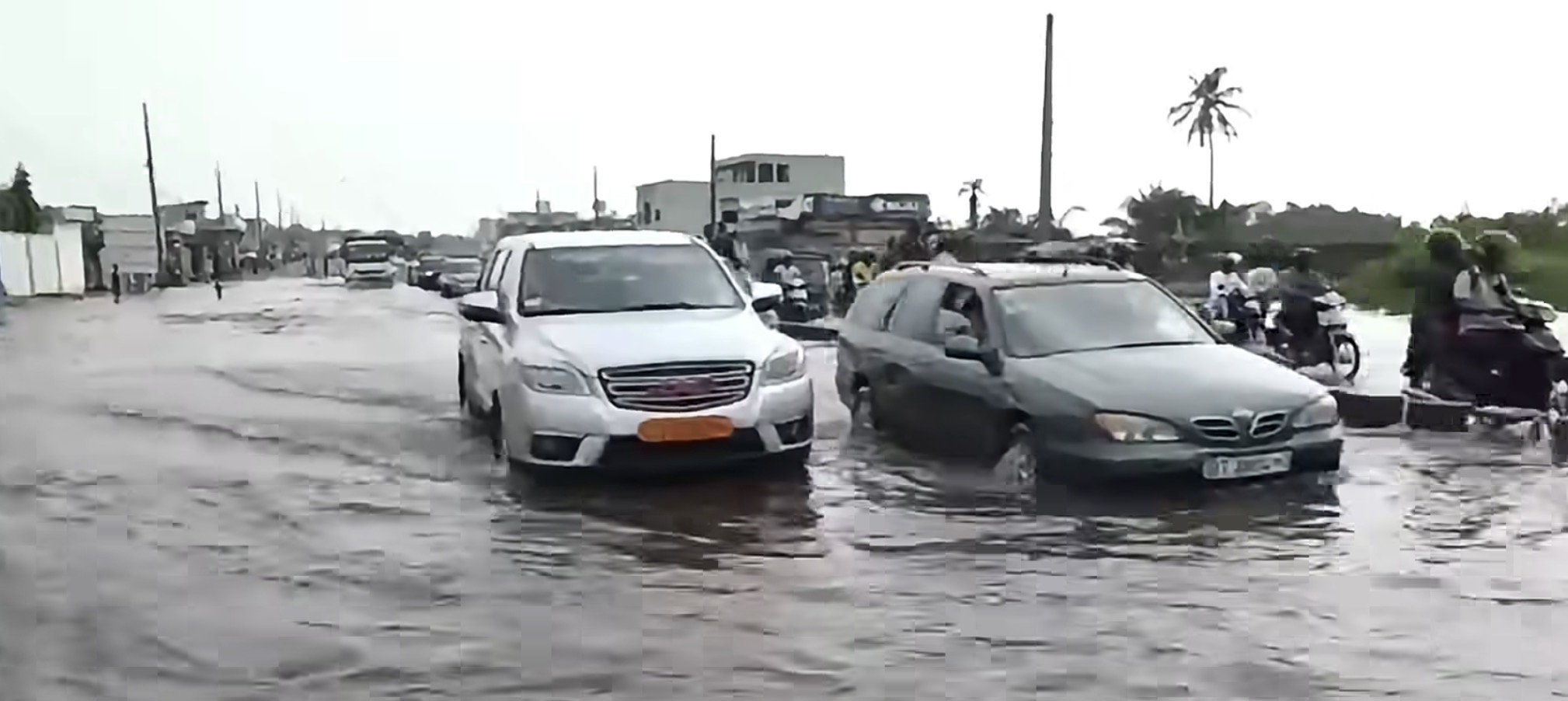 Voitures circulant dans une rue inondée avec des motos et des piétons sur le côté sous un ciel gris