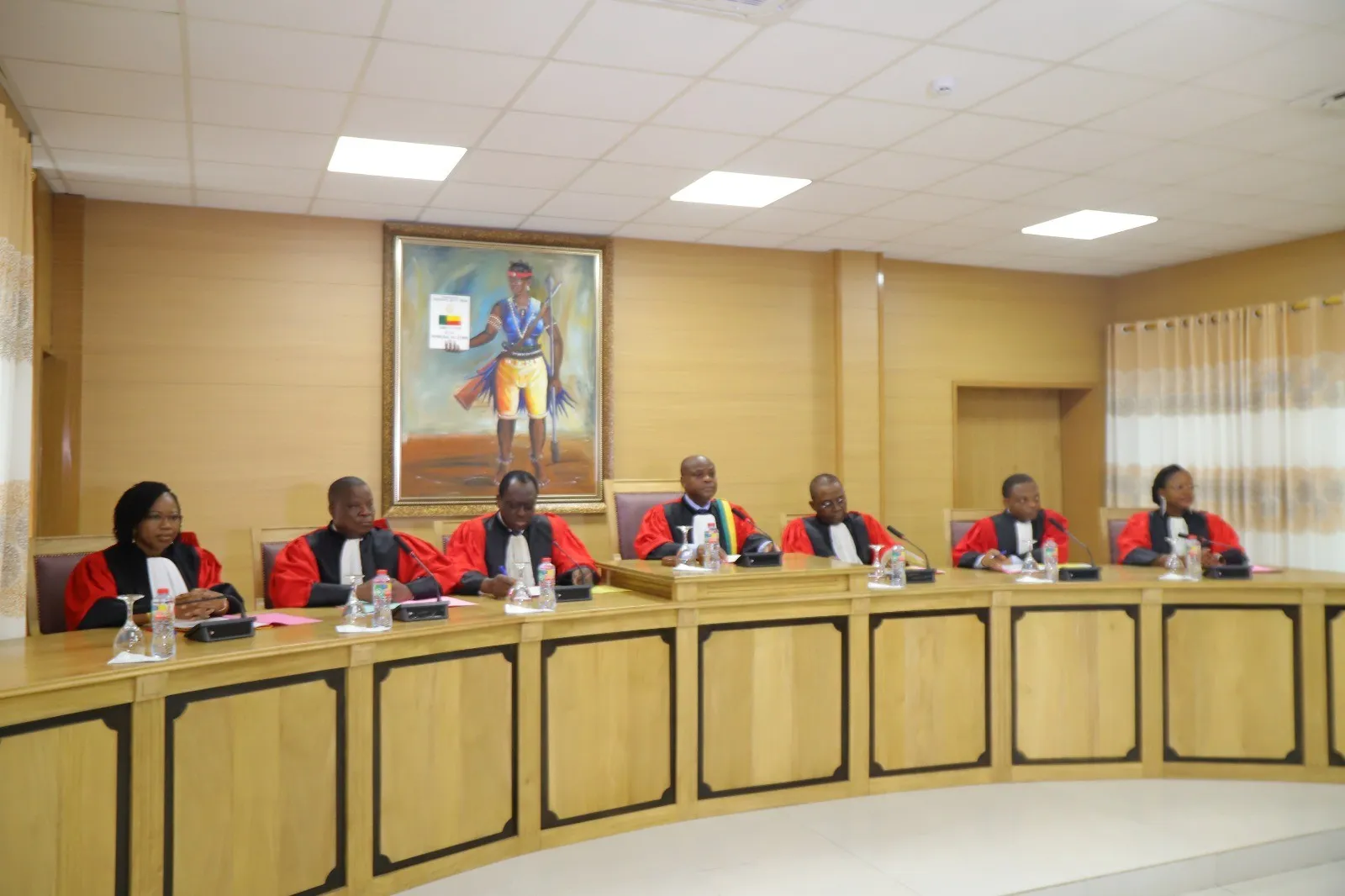 Sept personnes en robe rouge et noire assises derrière une table en bois dans une salle avec un tableau accroché au mur